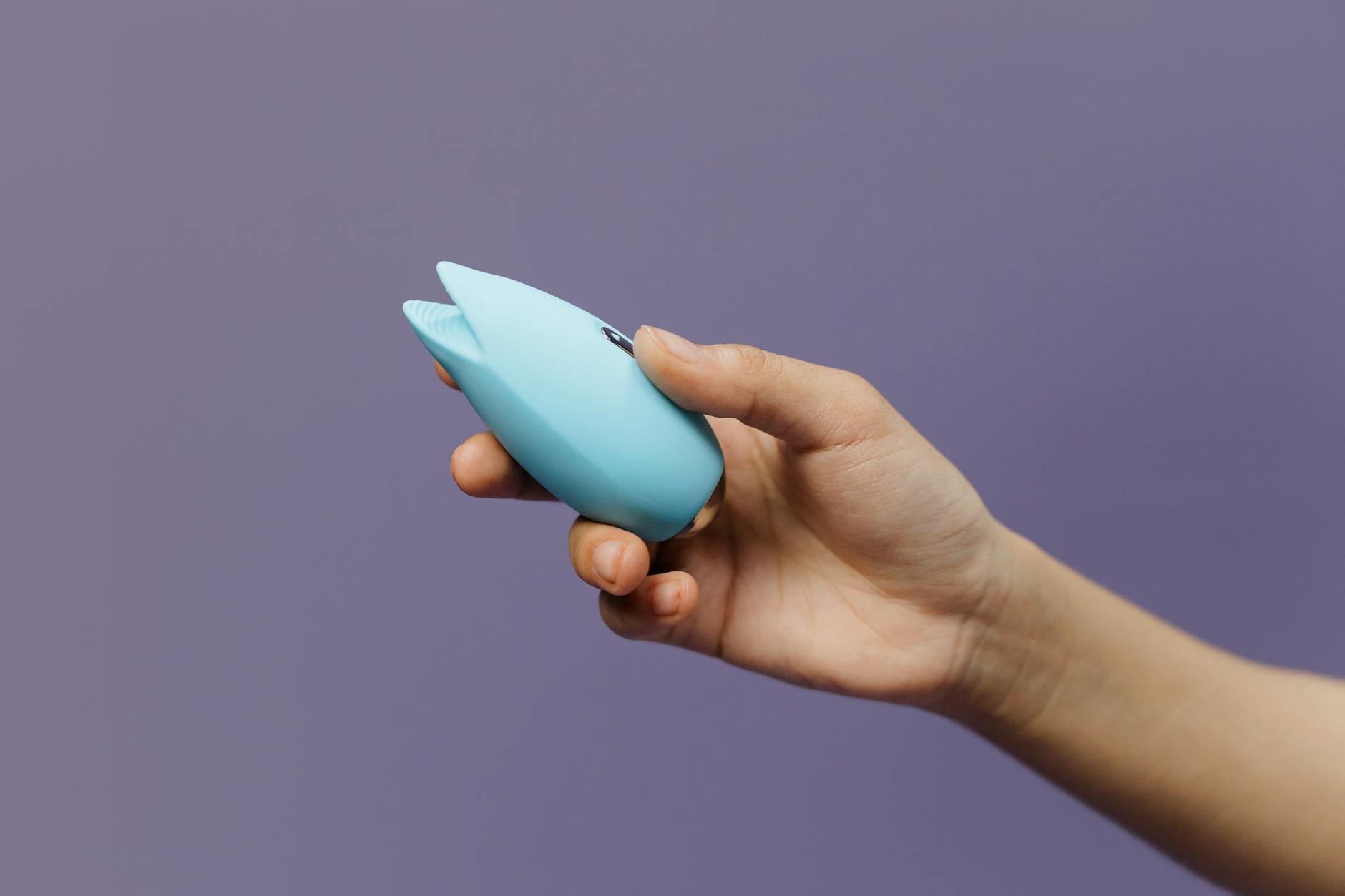 A blue silicone clitoral vibrator held in hand against a solid purple background, promoting self-love and intimacy.